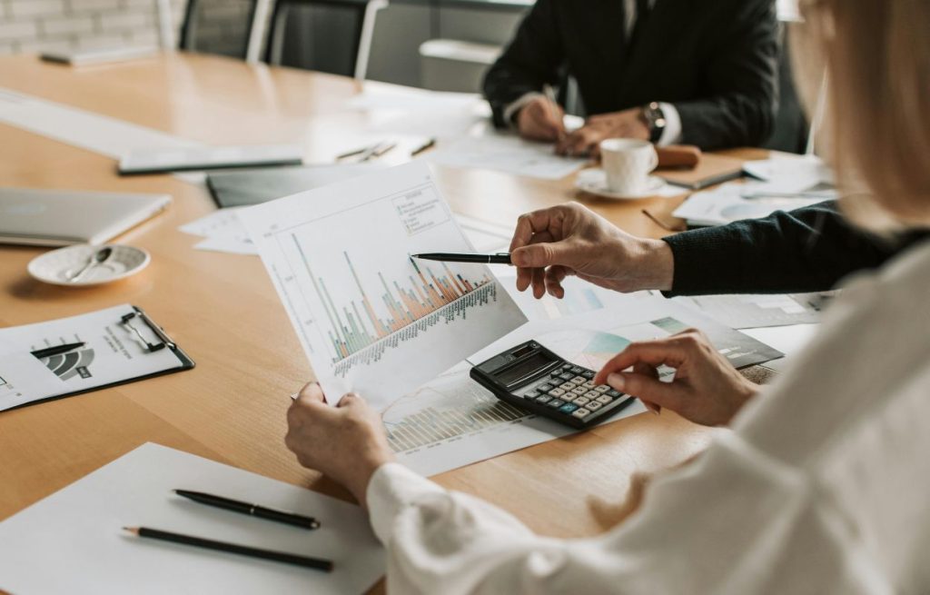 Pessoas ao redor de uma mesa com papéis, calculadora. Estão examinando gráficos.